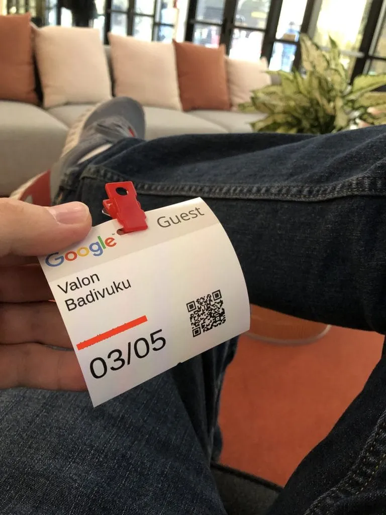 A Google guest badge reading "Valon Badivuku, 03/05" held in my hand over my knee, in a lounge with a sofa and cushions behind.