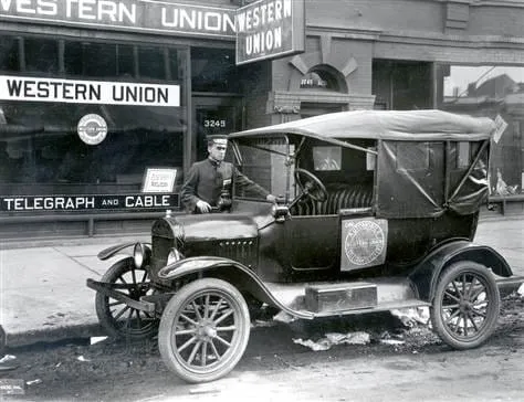 A black-and-white photograph of a man standing on the running board of an early Ford motor car parked outside a Western Union telegraph and cable office.