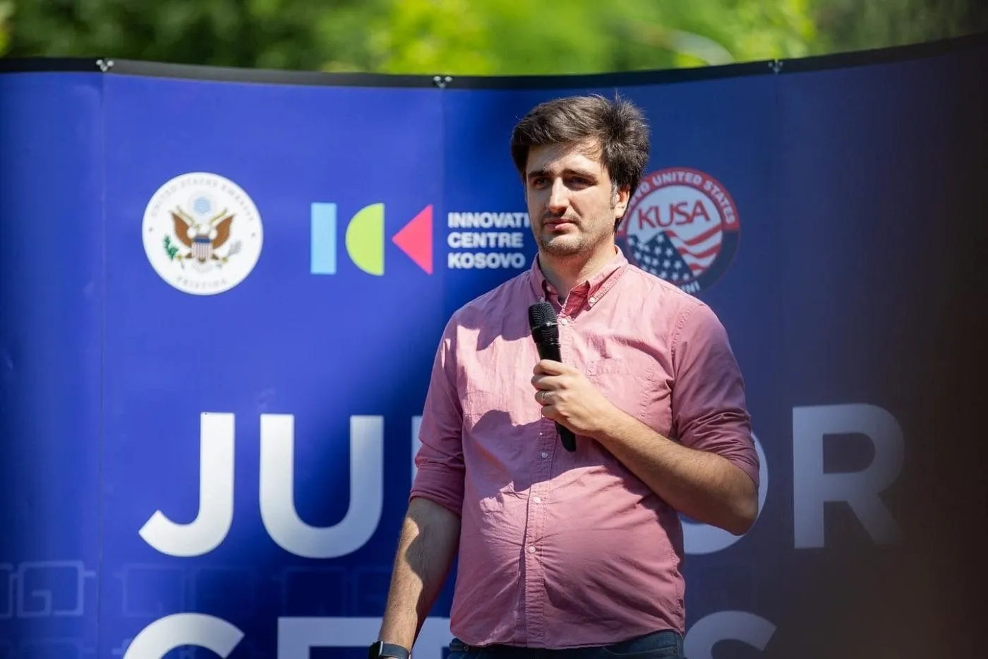 Me speaking into a handheld microphone at the JuniorGeeks graduation, in front of a blue backdrop carrying the seals of the U.S. Embassy Prishtina, the Innovation Centre Kosovo and KUSA.