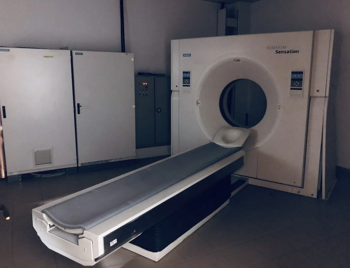 A Siemens Somaton Sensation CT scanner with its patient bed extended, standing in a bare room beside grey equipment cabinets.
