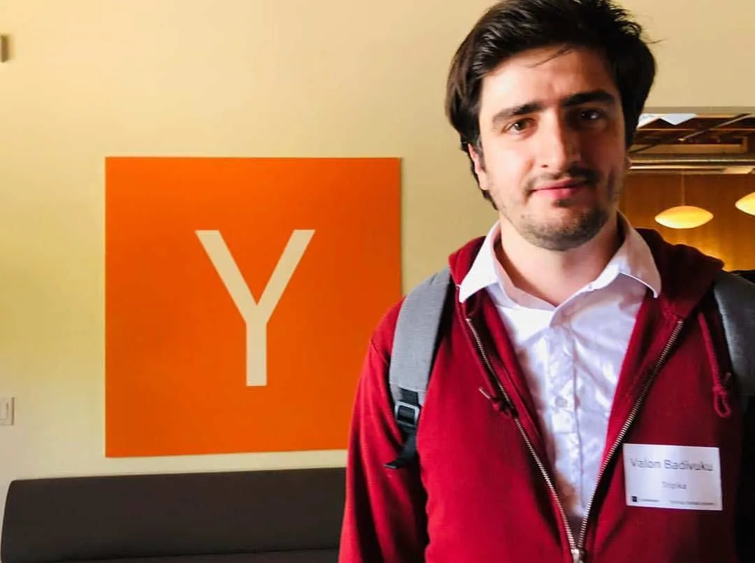 Me in a red hooded jacket and a Startup School name badge, standing in front of the orange Y Combinator logo on the wall.