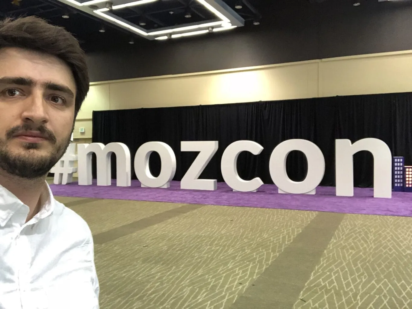 A selfie of me in the MozCon hall, in front of giant white letters spelling #mozcon on a purple carpet.