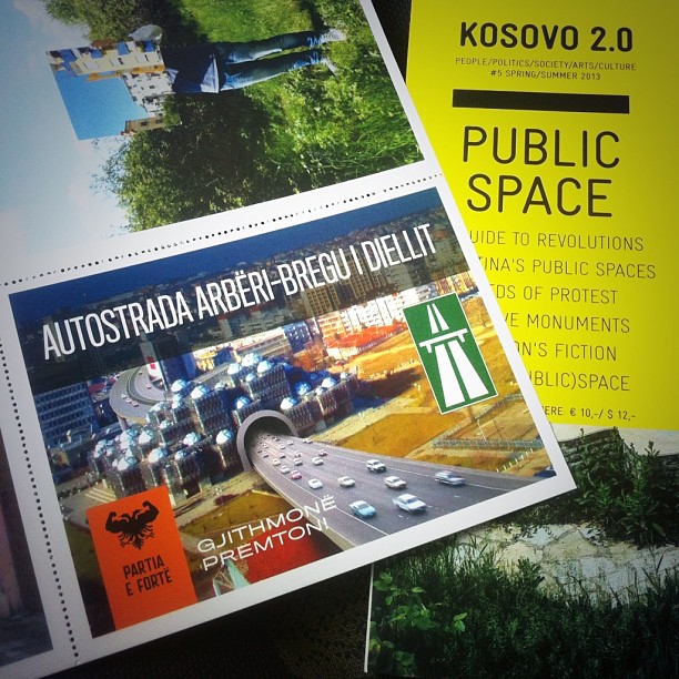 A Partia e Fortë postcard promising the "Autostrada Arbëri-Bregu i Diellit", showing a glowing multi-lane motorway over Prishtina with a green motorway sign and the slogan "Gjithmonë premtojmë", lying on top of Kosovo 2.0's Public Space issue.