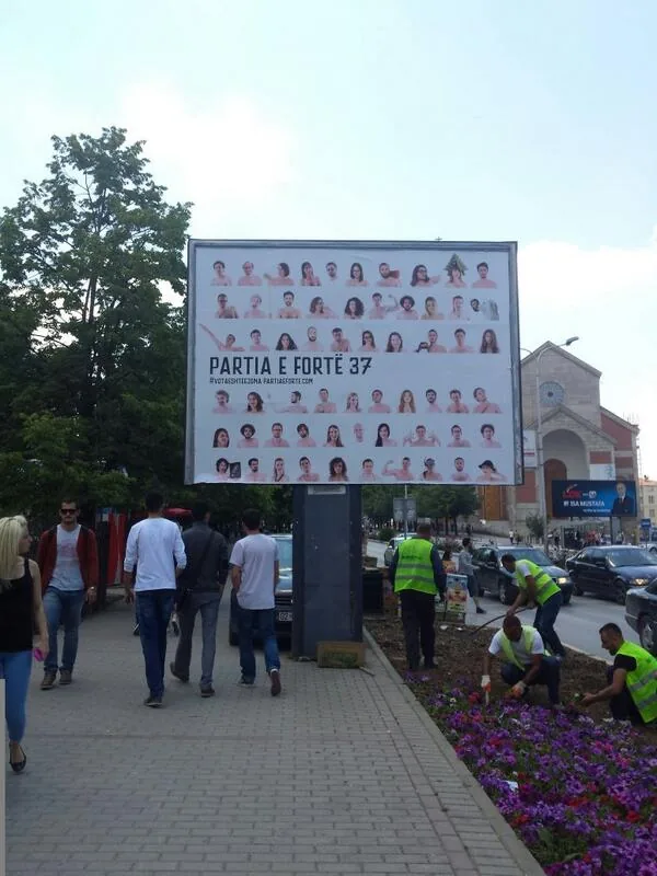 A Partia e Fortë billboard in downtown Prishtina showing the full grid of candidate portraits above the line "PARTIA E FORTË 37", with people walking past and workers planting flowers below it.