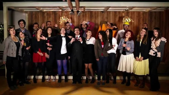 A still from the "Do të bëhem milioner" music video: about twenty people crowded together singing to camera, a man in a dark suit and red tie front and centre, and a pair of legs dangling from the ceiling above them.