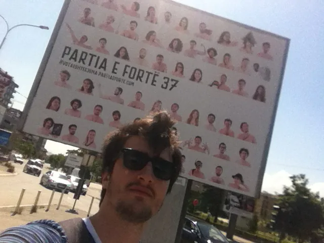 Me next to a Partia e Fortë street billboard in Prishtina, the whole grid of candidate faces behind me.