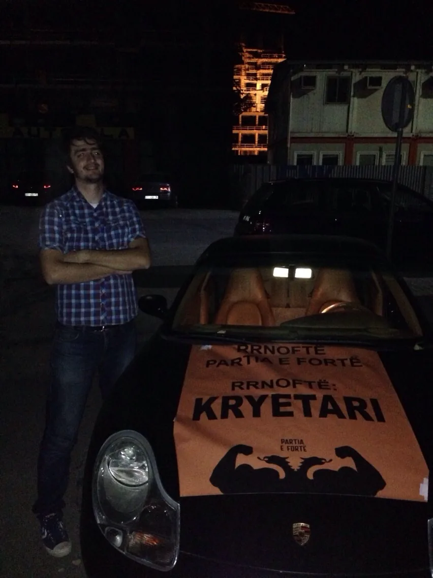 Me standing arms folded at night beside a black Porsche whose windscreen and bonnet are covered by an orange Partia e Fortë banner reading "Rrnoftë kryetari" (long live the leader).