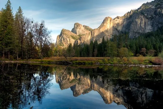 A granite mountain face and a line of evergreens mirrored in the still water of a valley river at dusk.