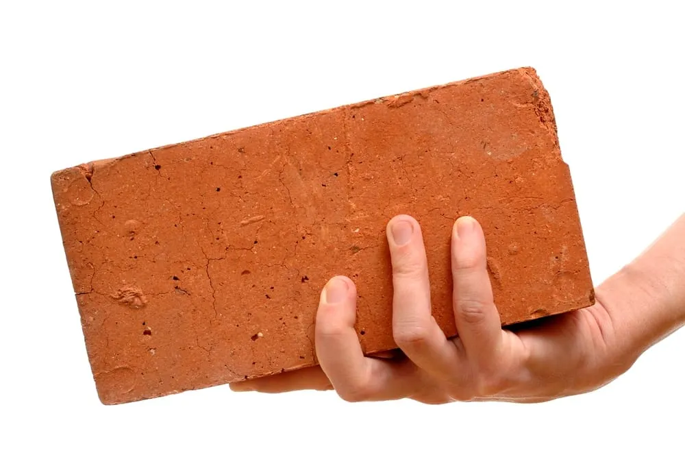 A hand holding up a single red clay brick against a white background.
